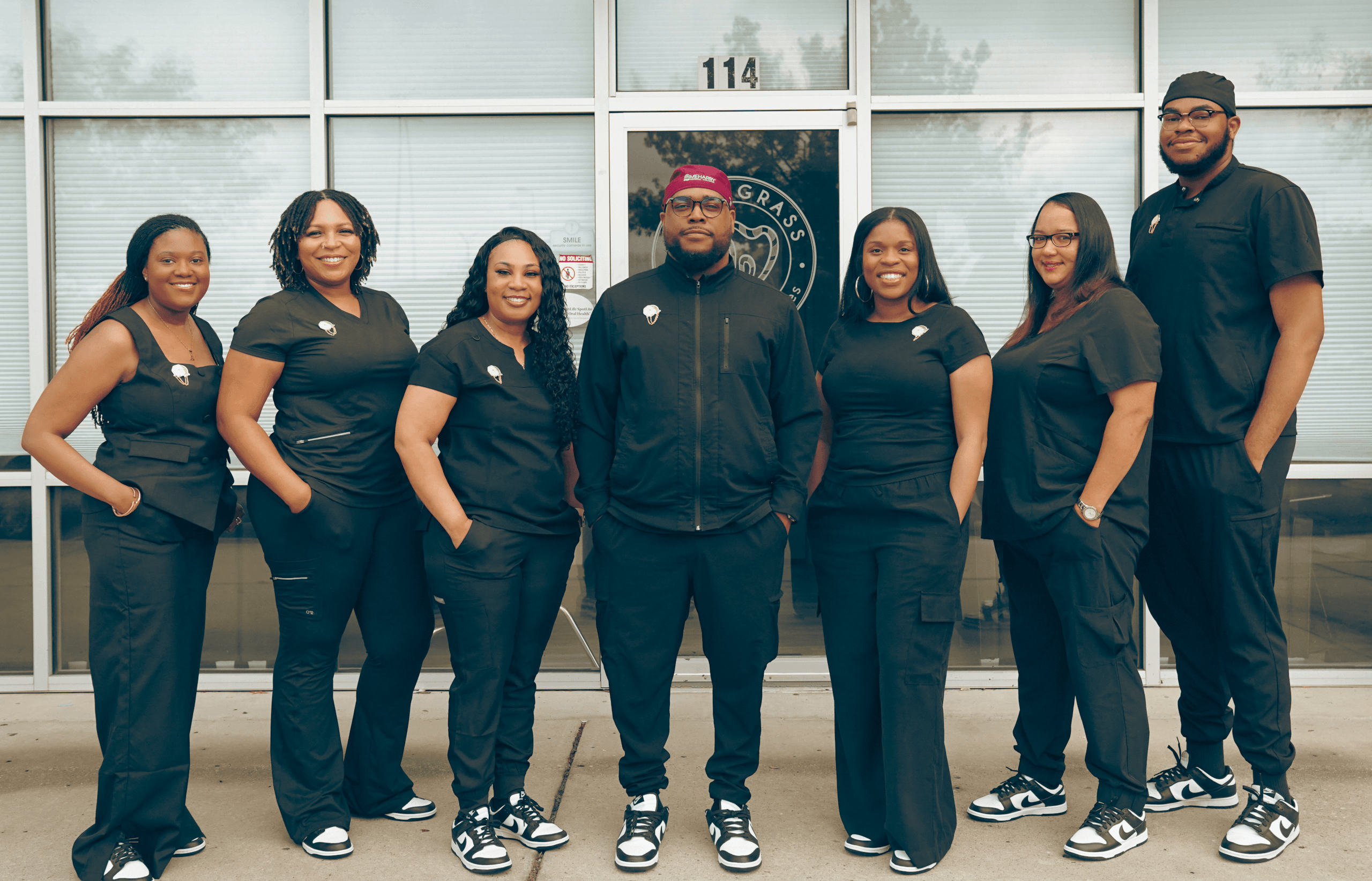 Shawn Mitchell, DDS, with team in front of clinic – SweetGrass Dental Associates Dr. Shawn Mitchell, DDS, standing with six smiling staff members outside the SweetGrass Dental Associates office, all dressed in matching black scrubs and sneakers, posed confidently in front of a glass door marked with the clinic logo – SweetGrass Dental Associates