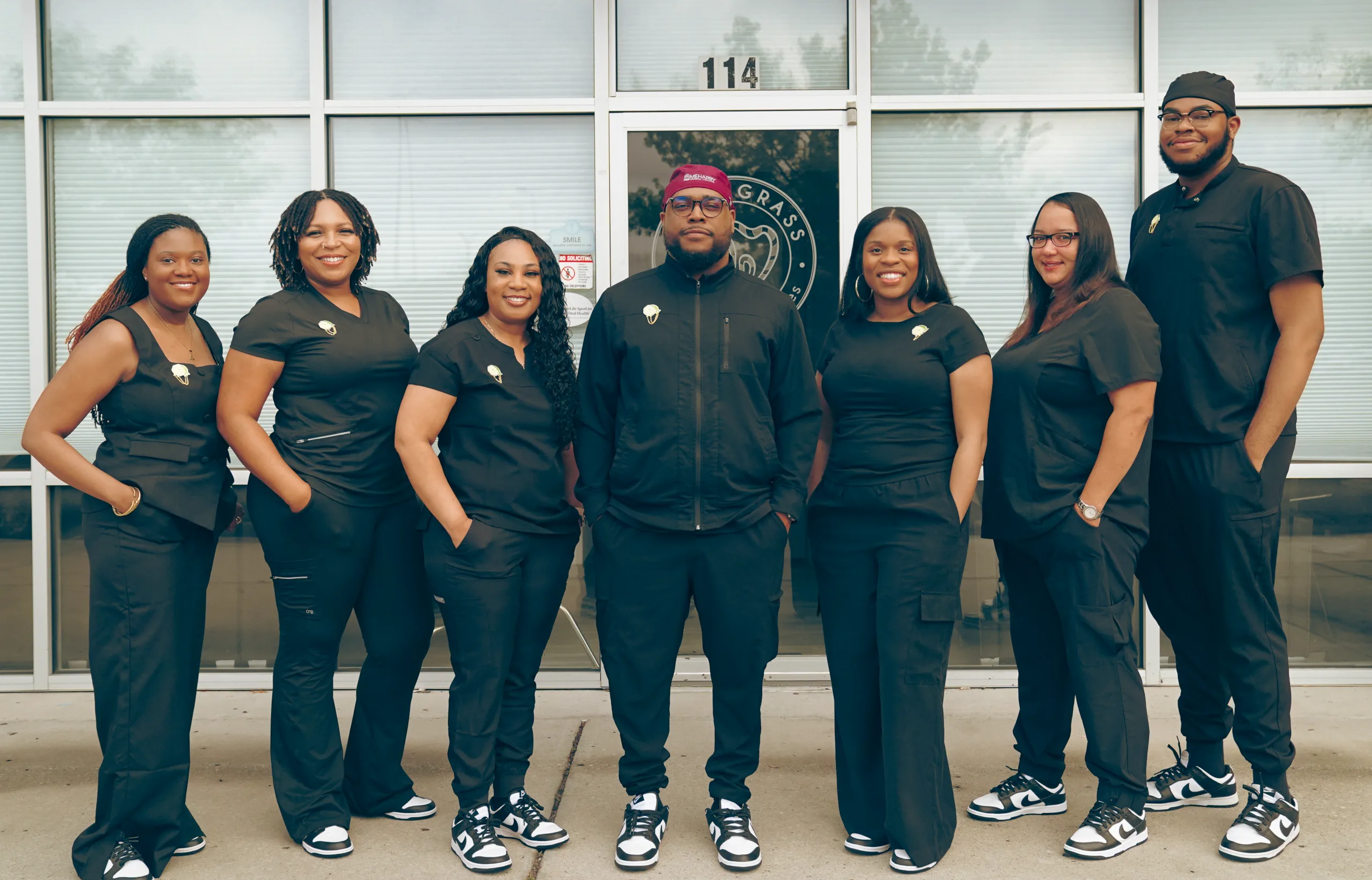 Dr. Shawn Mitchell, DDS, standing with six smiling staff members outside the SweetGrass Dental Associates office, all dressed in matching black scrubs and sneakers, posed confidently in front of a glass door marked with the clinic logo – SweetGrass Dental Associates